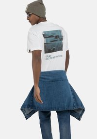 Camiseta blanca con un gráfico impreso en la parte de atrás, combinada con unos jeans de mezclilla azules y un envoltorio de mezclilla. Un gorro gris y gafas de sol completan el look.