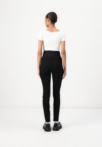 Jeans negros de cintura alta con un corte ajustado, que cuentan con bolsillos traseros y una textura de tela suave, combinados con una blusa blanca ajustada y zapatos negros.