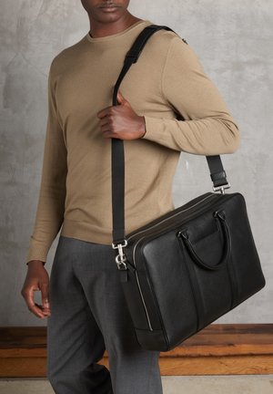 Sac messenger en cuir noir avec deux poignées et une bandoulière réglable. Texture lisse, fermeture éclaire et coutures verticales simples.