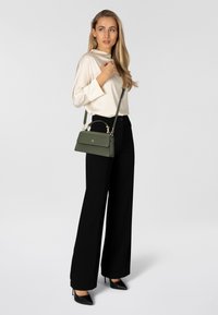 Borsa a mano in pelle verde con design strutturato, manico superiore e tracolla staccabile. Abbinata a una camicia bianca in raso e pantaloni neri a gamba larga.