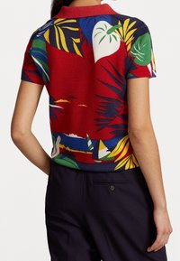 Kortärmad färgglad skjorta med tropiska blad- och segelbåtsmönster, bärs med mörka byxor sett bakifrån.