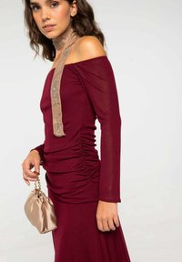 Robe bordeaux décolletée avec des manches longues, texture froncée, et un accessoire de cou en écharpe beige ornée. Tient un sac doré en satin.