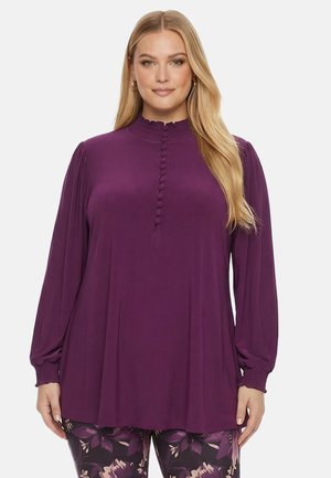 Frau trägt eine langärmelige lila Bluse mit stoffbezogenen Knöpfen und Blumenmuster-Hose und steht vor einem einfarbigen Hintergrund.