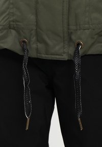 Veste verte à surface texturée, dotée de cordons noirs avec des éléments dorés. Pantalon noir à texture lisse visible en dessous.
