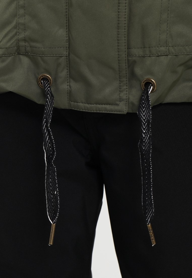 Veste verte à surface texturée, dotée de cordons noirs avec des éléments dorés. Pantalon noir à texture lisse visible en dessous.