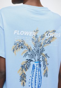 Φωτεινό γαλάζιο βαμβακερό μπλουζάκι με σχέδιο ενός ριγέ αγγείου με κίτρινες και πρά�σινες λουλουδένιες εικόνες και το κείμενο «ΛΑΤΡΕΙΣ ΤΩΝ ΛΟΥΛΟΥΔΙΩΝ».