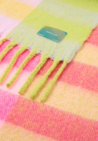 Třásňový šátek v jasně růžovém a zeleném kostkovaném vzoru, vyrobený z měkkého materiálu. Obsahuje štítek s nápisem "ARCTIC AIR", který zdůrazňuje jeho živé barvy.