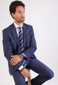 Marineblauw pak met een slim fit ontwerp, een wit overhemd en een gestreepte stropdas. Accessoires zijn een horloge en armbanden om de pols.