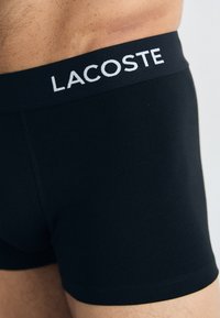 Black cotton boxer shorts with a wide elastic waistband featuring "LACOSTE" logo in white. Smooth texture, fitted design, mid-thigh length.