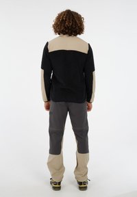 Persona con cabello rizado que lleva una chaqueta de forro polar negra y beige, pantalones bicolores en gris y beige, y zapatos de exterior negros, de pie mirando hacia fuera.
