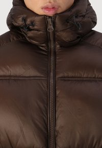 Chaqueta acolchada marrón con un cuello alto, cremallera frontal y cordones ajustables. Acabado texturizado con paneles acolchados para aislamiento.