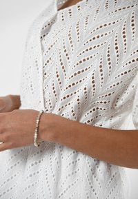 Een close-up van de hand van een persoon die een delicaat armband draagt, met een witte, ingewikkeld geprinte stoftop met bladachtige ontwerpen.