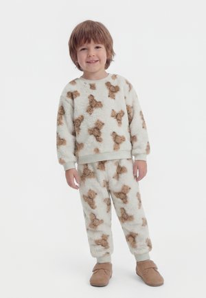 Ungt barn som står, iført hvitt, mykt pysjamasett med brune hundemønstre og lysebrune sko, smilende mot kamera.