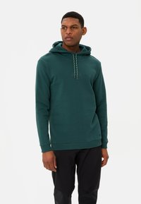 Dunkelgrüne Kapuzenjacke aus weichem Material, mit verstellbarem Kapuzenzug und gerippten Bündchen, kombiniert mit schwarzen Hosen.