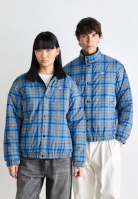 Padded jackets with blue plaid pattern, black accents, button closure, and a collar. One has a fleece collar; both feature elastic cuffs.