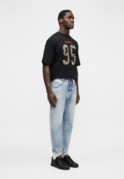 Homme debout portant un t-shirt noir avec "95" en imprimé léopard, un jean bleu clair, des chaussettes blanches et des baskets noires sur un fond blanc.