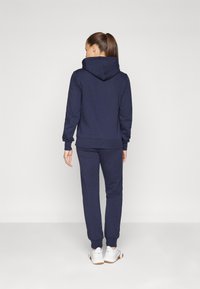 Ensemble de sweat à capuche et de pantalon de jogging bleu marine, fabriqué dans un matériau doux, avec une grande capuche et des poignets côtelés. Le mannequin est de dos, portant des baskets blanches.