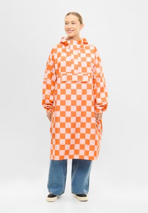 Kobieta w pomarańczowo-różowym długim płaszczu przeciwdeszczowym w kratę z kapturem, w połączeniu ze szerokimi niebieskimi dżinsami i czarno-białymi butami.