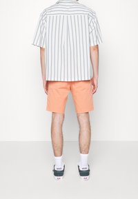 Chemise à manches courtes rayée blanche et bleue, associée à un short orange. Chaussures : baskets vert foncé et blanches avec un logo visible.