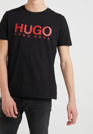 Jonge persoon die een zwart T-shirt draagt met grote rode "HUGO BOSS" tekst op de voorkant, gecombineerd met donkere jeans tegen een neutrale achtergrond.