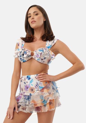 Donna che indossa un crop top colorato a fiori con volant e dettaglio ad anello, abbinato a una minigonna a strati su sfondo semplice.