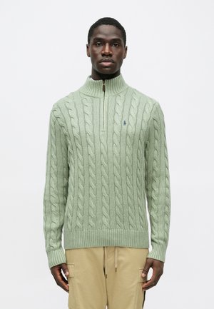 Vihreä kaapeli-neulepusero, jossa on korkea kaulus ja neljännesvetoketju. Ribatut hihansuut ja helma. Rinnassa pieni logo.