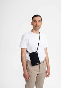 Zwarte stoffen schoudertas met verstelbare band en logo. Rectangulaire vorm, minimalistisch ontwerp, voorzien van een ritsluiting en twee oogjes.