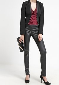 Blazer negro ajustado con un top burdeos de escote en V profundo, leggings negros brillantes y zapatos de tacón altos negros. Sosteniendo un bolso de mano negro con un acento dorado.