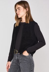 Gilet a maglia nera con una finitura morbida e testurizzata, caratterizzato da scollatura rotonda e senza chiusure, indossato sopra una camicia nera a maniche lunghe e jeans grigi.