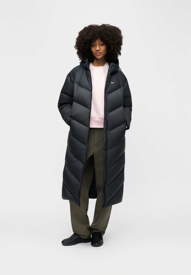 Frau steht mit den Händen in den Taschen, trägt einen langen schwarzen Steppmantel, ein hellrosa Sweatshirt, olivgrüne Hosen und schwarze Turnschuhe.