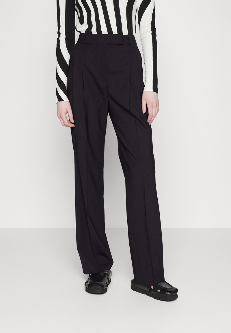Personne portant un pantalon noir ajusté, des sandales noires épaisses à bout ouvert, et un haut à manches longues à rayures verticales noires et blanches.