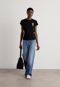 Black cotton t-shirt with a star graphic, paired with light blue wide-leg jeans and white sneakers. Model carries a black bag.