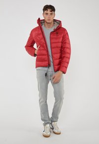 Giacca a vento rossa con cappuccio, chiusura a zip e polsini elasticizzati. Indossata sopra una t-shirt grigia, abbinata a jeans grigi chiari e sneakers bianche.