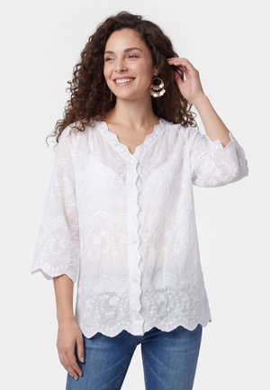 Vrouw met krullend haar die lacht, een witte geborduurde blouse met knopen en geschulpte randen en een blauwe spijkerbroek draagt, terwijl ze haar haar aanraakt.