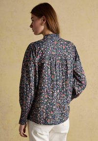 Sieviete, tērpusies tumši zilu ziedu blūzē ar garām pūkainām piedurknēm un smockētu aproču daļu, kopā ar baltām biksēm, stāvot pretī bēšam fona.