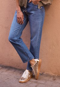 Guldmetalliska sneakers med vita detaljer och snören. Blå denimbyxor med rak passform. Bakgrunden har en texturerad vägg.