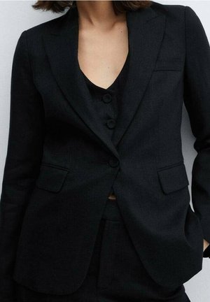 Vrouw draagt een zwarte, op maat gemaakte blazer met bijpassend gilet met knopen en zwarte broek, staand met één hand in de zak.