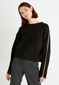 Maglione nero a costine con maniche ampie e una striscia bianca lungo un lato. Abbinato a pantaloni grigi su misura.