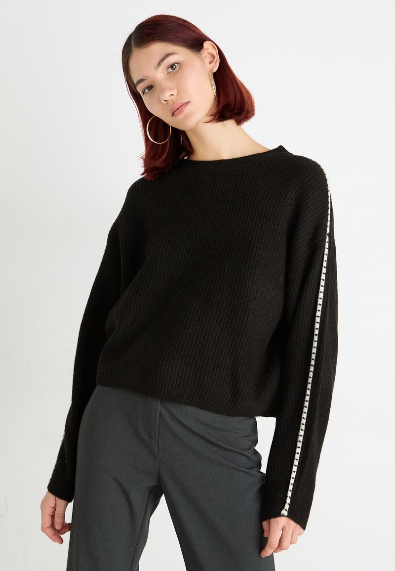 Maglione nero a costine con maniche ampie e una striscia bianca lungo un lato. Abbinato a pantaloni grigi su misura.