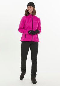 Fleece-takki kirkkaan vaaleanpunaisena, korkealla kauluksella, täysipitkällä vetoketjulla ja mustilla yksityiskohdilla; yhdessä mustien housujen ja käsineiden kanssa.