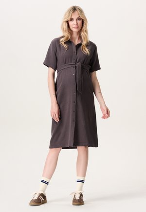 Grijze jurk met knopen gemaakt van gestructureerde stof en een strikceintuur in de taille. Korte mouwen en knie lengte, gecombineerd met witte sokken en bruine schoenen.