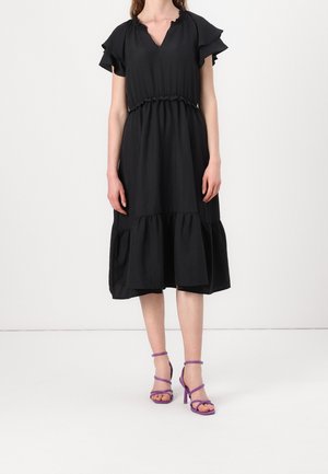 Femme portant une robe midi noire avec des manches et un ourlet à volants, associée à des sandales à talons hauts violettes à lanières, debout devant un fond blanc.