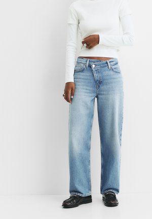 Hellblaue Denim-Jeans mit weit geschnittenem Bein, ausgestattet mit einem Knopfverschluss, Gürtelschlaufen und minimaler Ausbleichung. Kombiniert mit einem weißen, figurbetonten Langarmtop.