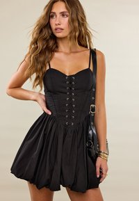 Femme aux longs cheveux ondulés portant une robe corset noire à lacets et un sac à épaule noir, posant avec une main sur la hanche.