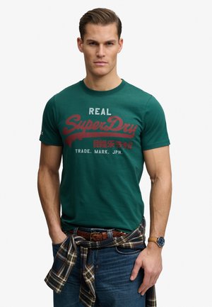 Mann trägt grünes T-Shirt mit "SuperDry"-Aufdruck, blaue Jeans, kariertes Hemd um die Taille gebunden und Armbanduhr, steht mit Händen in den Taschen.