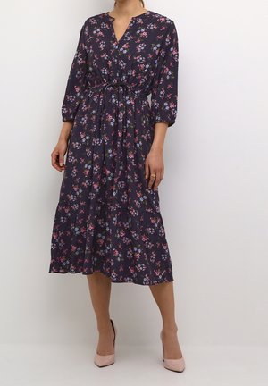 Kvinde iført en navy-midi-kjole med pink og lilla blomsterprint, 3/4 ærmer, indsnøret talje og lyserøde høje hæle.
