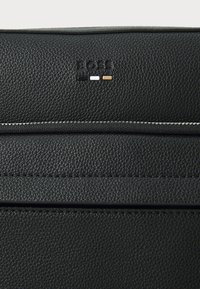 Borsa in pelle nera con texture e logo in rilievo. Presenta accenti bianchi e marroni in contrasto, cuciture argentate eleganti e un design minimalista.