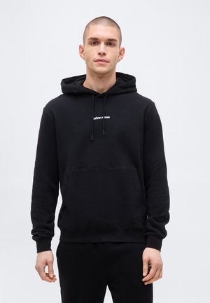 Junger Mann mit kurzen Haaren, trägt einen schwarzen Kapuzenpullover mit dem Schriftzug "BJÖRN BORG", steht vor einem schlichten hellgrauen Hintergrund.