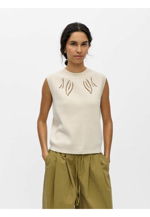 Kvinde iført en ærmeløs beige top med udskæringer i form af blade og højtaljede løse olivengrønne bukser med hænderne i lommerne.