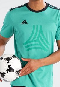 Turkusowa koszulka sportowa z czarnym kołnierzykiem i paskami, z geometrycznym wzorem z przodu. Czarno-biała piłka nożna trzymana w dłoni.